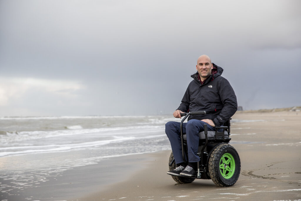 Man in een Bi-Go balansrolstoel rijdt langs de kustlijn op het strand.