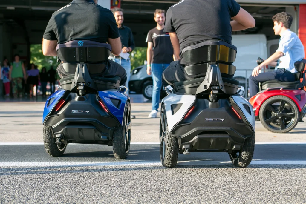 Twee gebruikers rijden samen buiten op Genny zero-balansrolstoelen, terwijl anderen in de achtergrond toekijken.