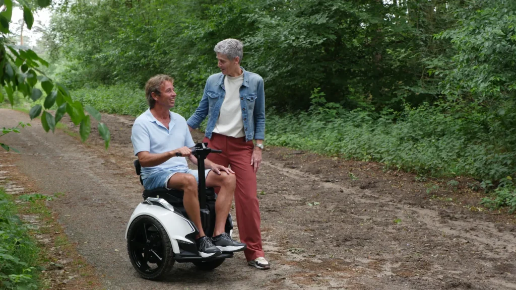 Man in een Genny Zero balansrolstoel met een begeleider naast zich op een bosachtig pad.
