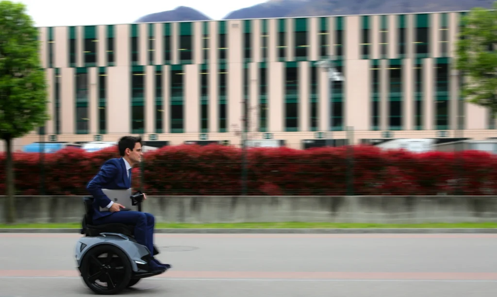 Man in pak rijdt met een Genny Zero balansrolstoel over straat langs een modern kantoorgebouw.